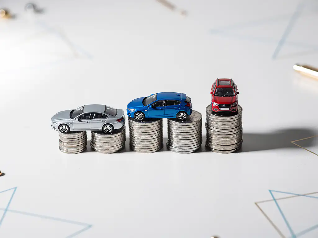 Drie miniatuur auto's op stapels munten tonen kostenvergelijking met zilveren sedan, blauwe hatchback en rode SUV