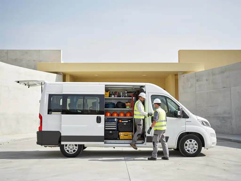 Witte minibus met bouwgereedschap en helmen bij moderne bouwplaatsingang, twee werknemers stappen in, zacht daglicht.