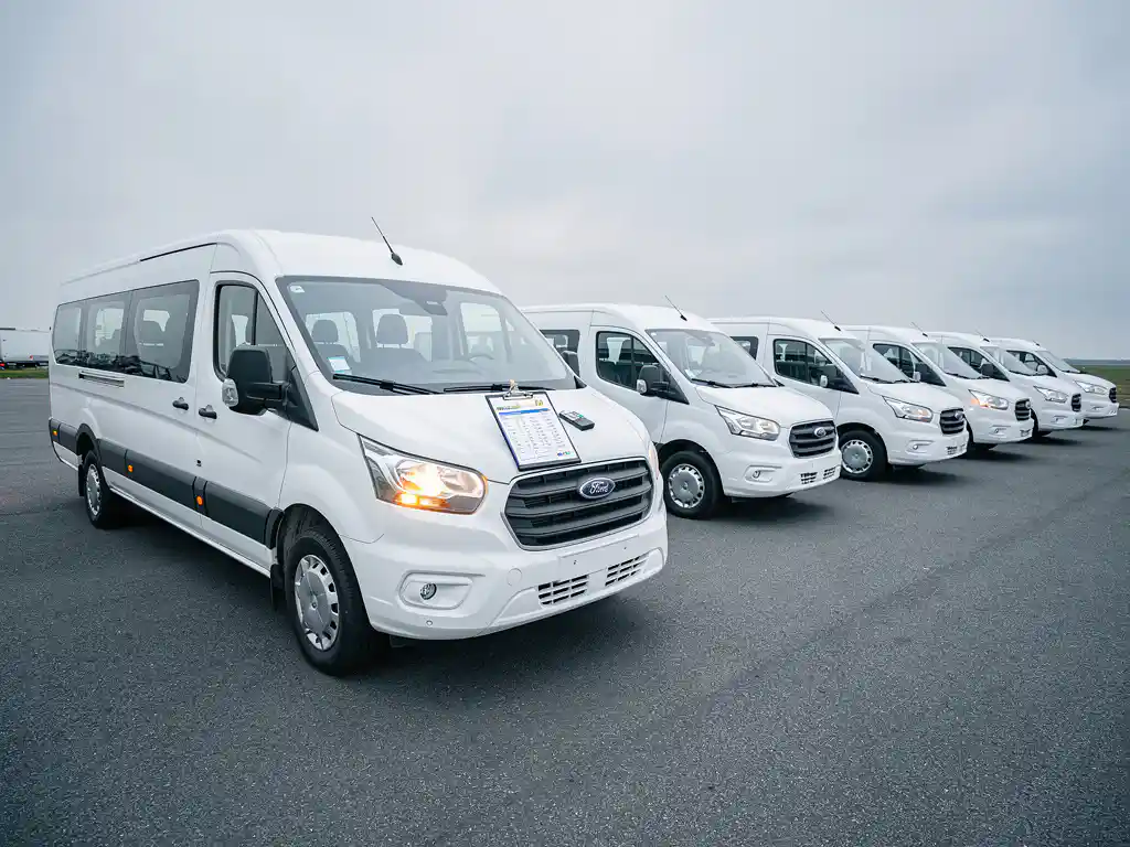 Witte passagiersminibus geparkeerd naast een vloot identieke huurbusjes op een asfaltterrein, met een klembord op de motorkap.