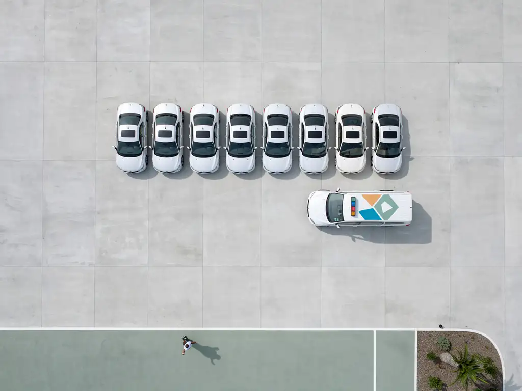 Witte auto's in geometrische rij naast een gemarkeerd zorgvoertuig, luchtfoto op betonnen ondergrond in minimalistische compositie.