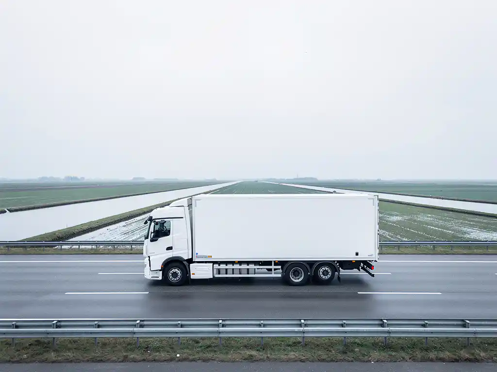 Witte vrachtwagen op Nederlandse snelweg met vlak polderlandschap en bewolkte lucht op de achtergrond.