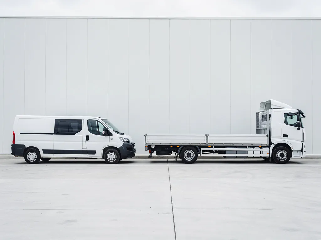 Witte bestelwagen geparkeerd naast een grote flatbed vrachtwagen op betonnen ondergrond, gefotografeerd in zacht daglicht.