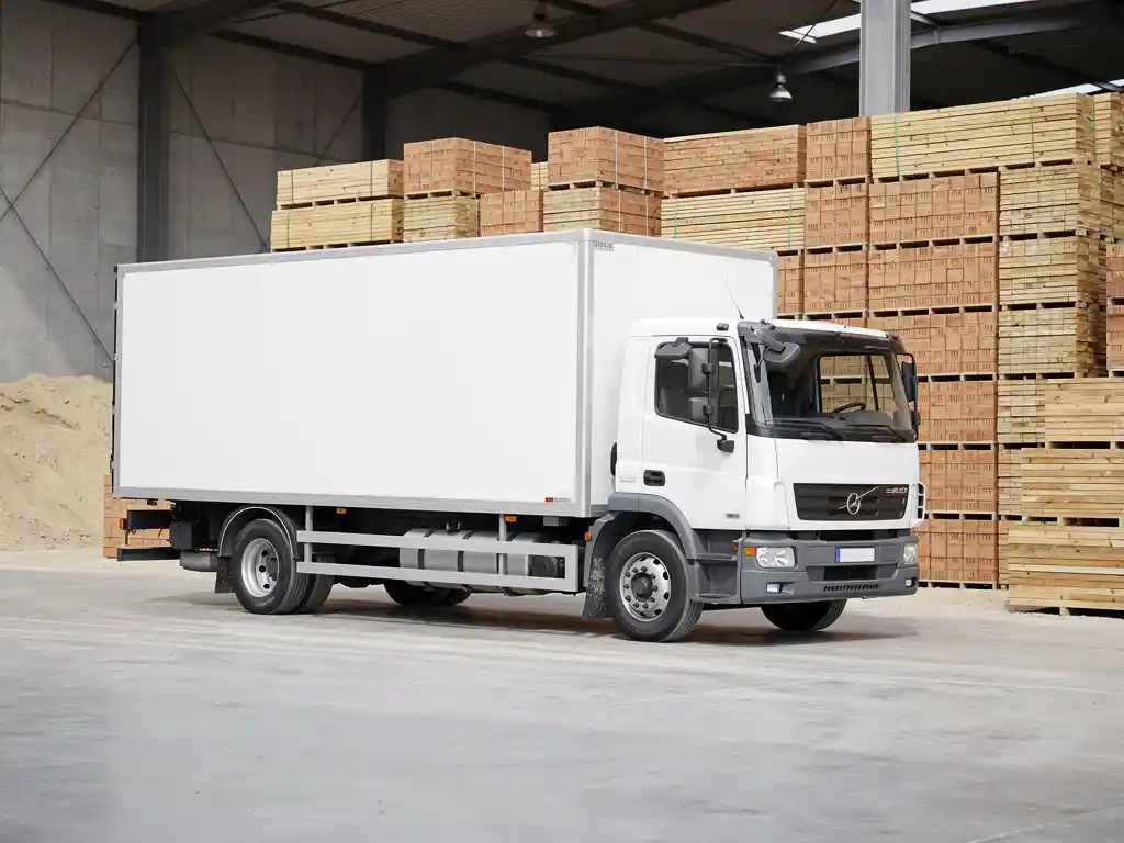 Witte vrachtwagen geparkeerd op betonnen ondergrond bij bouwplaats, met stapel bouwmaterialen op de achtergrond.
