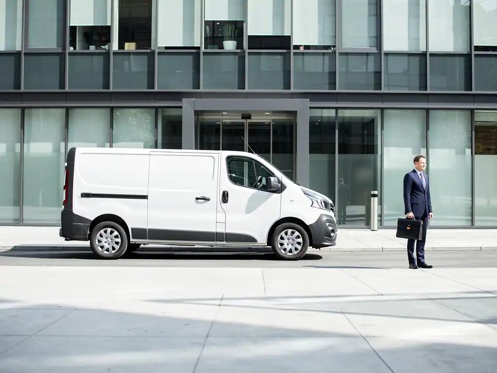 Witte bezorgbus arriveert bij modern glazen kantoorgebouw, professional met aktetas wacht op stoep in middaglicht.