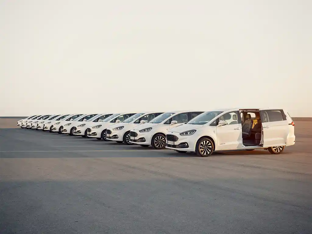 Witte minibussen in diagonale rij geparkeerd op een open terrein, één busje naar voren getrokken met open schuifdeur in gouden uurtje licht.