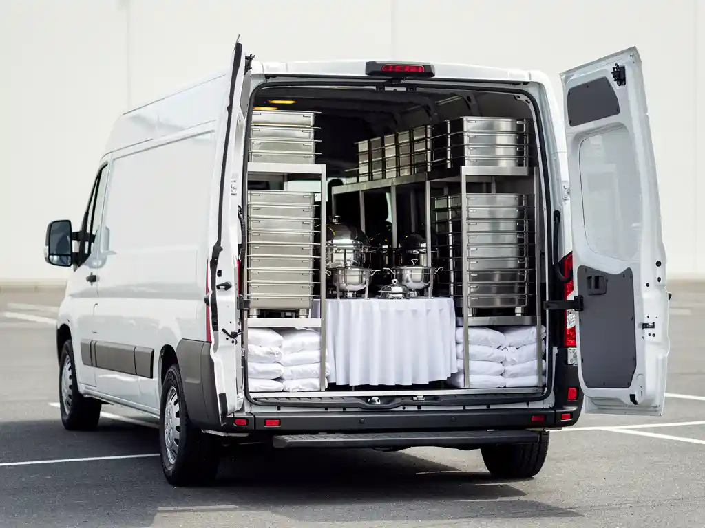 Witte cateringbus met open achterdeuren vol netjes gestapelde zilveren serveerschalen, tafelkleden en roestvrijstalen apparatuur.